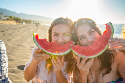 Two women eating watermelon and looking at the camera. They are smiling and happy. They are making smiley faces with their watermelons. Beach can be seen in the background.
