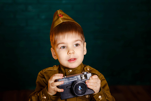 Boy in russian military uniform with camera. Child war correspondent during the Second World War. Photographer with camera in his hands. Military reconstruction. Soldier with film camera