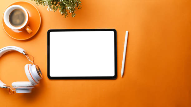 Mock up digital table with blank screen, coffee cup and headphone on orange background.