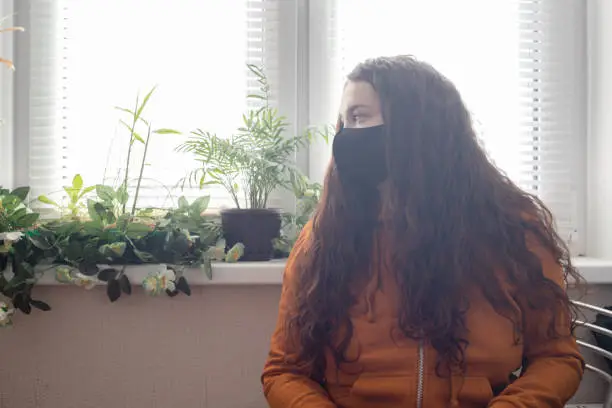 Photo of Red-haired curly girl in an orange jacket and black mask on her face sits near the window