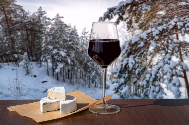 Glass of wine with brie cheese on  winter forest landscape background. View of covered in snow pine and fir trees. stock photo