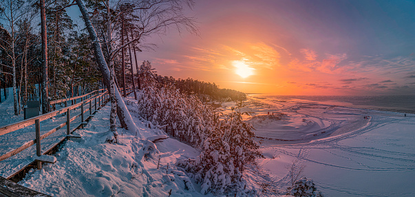 Colorful sunset over snowy sea coast and pine forest with wooden boardwalk path. Beautiful view of Baltic sea coast with snow-covered fir, spruce and pine trees and footpath for relaxing walk.