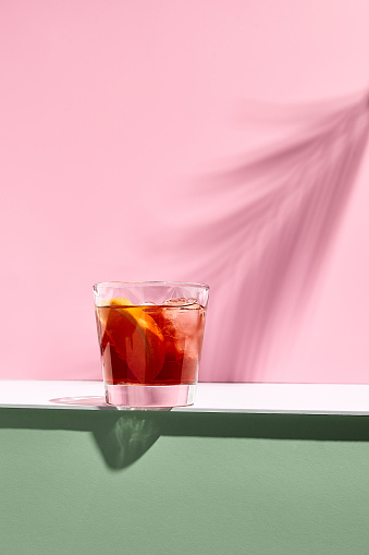 Negroni cocktail over pink background. Drink in rox glass in daylight with palm leaf hard shadow. Summer, tropical, fresh drink concept.