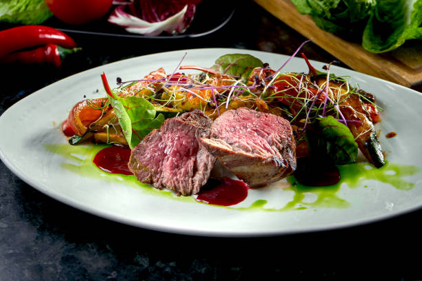 istock Medium rare beef medallions with vegetable garnish, served on a white plate. Dark marble background. Barbecue meat, restaurant food. Tasty steak 1305031589