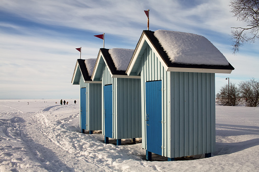 Three old beach huts stand empty on a beautiful winter day. People are walking on the ice and not going swimming.