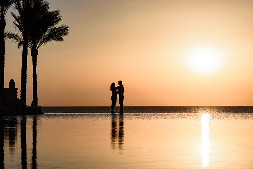 Silhouette of an affectionate couple standing on the edge of a swimming pool at sunset. Copy space.