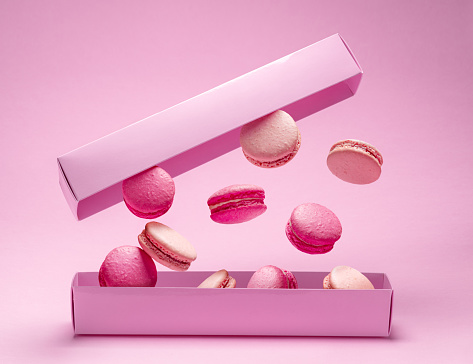 Colorful sweet macarons or macaroons, flavored cookies floating in the air above the paper box. Pink background.