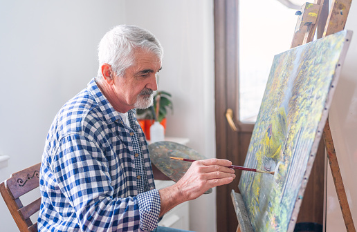 Senior man painting on a canvas seated on a chair at home. Senior artist working in concentration.