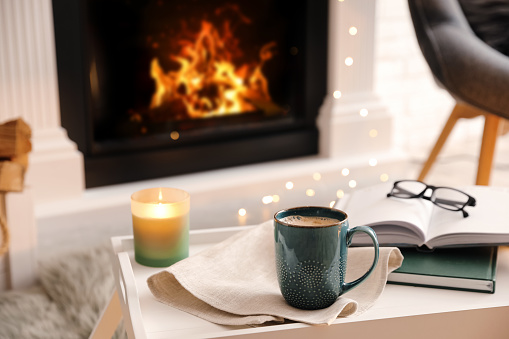Cup of coffee, burning candle and books on tray near fireplace indoors. Cozy atmosphere