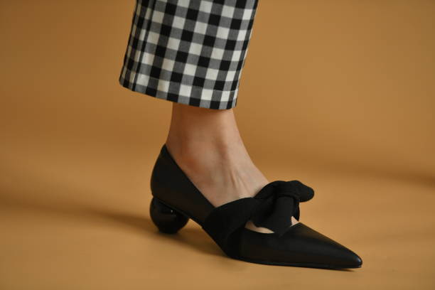istock Stylish female shoe with sharp sock, unique sphere heel and a cloth bow on top. Beautiful leg ankle and plaid pants trews. Natural colors and shadow, isolated on brown background. Fashion concept. 1305042035