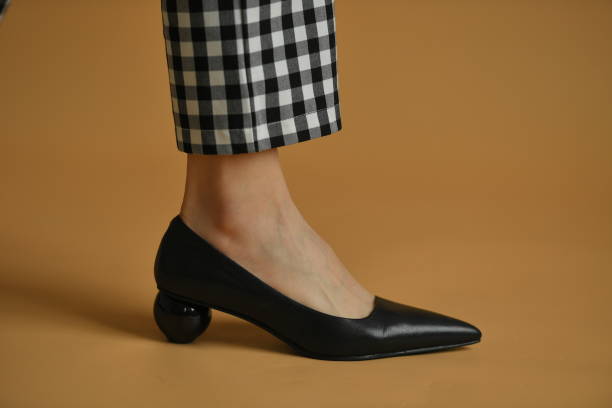istock Stylish female shoe with sharp sock, unique sphere heel and a cloth bow on top. Beautiful leg ankle and plaid pants trews. Natural colors and shadow, isolated on brown background. Fashion concept. 1305042141