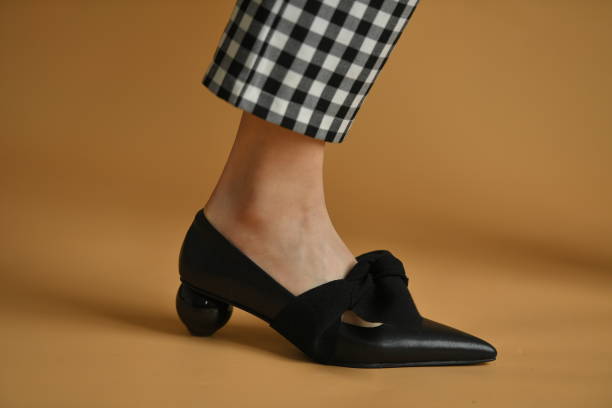 istock Stylish female shoe with sharp sock, unique sphere heel and a cloth bow on top. Beautiful leg ankle and plaid pants trews. Natural colors and shadow, isolated on brown background. Fashion concept. 1305042925