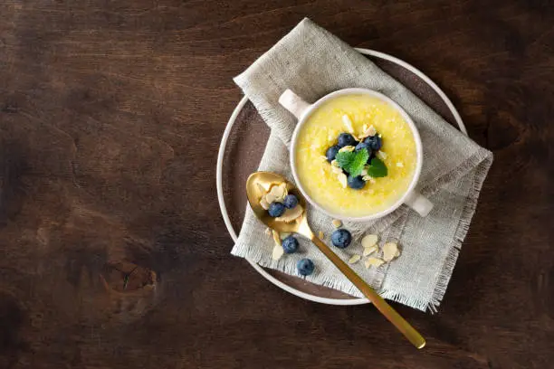 Polenta dish with blueberries and almonds. Corn porridge in a ceramic plate on the brown wooden kitchen table. Cream-soup with polenta. Healthy Vegetarian food