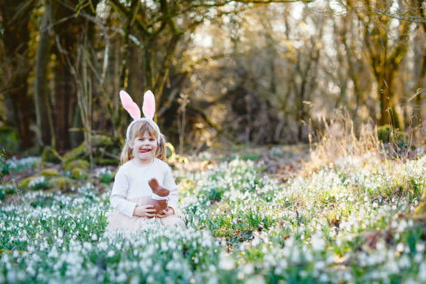 entzückende kleine mädchen mit osterhasen ohren essen chocholate figur im frühlingswald am sonnigen tag, im freien. nettes glückliches kind mit vielen schneeglöckchenblumen. frühlingshaftes, christliches urlaubskonzept. - bunny girl stock-fotos und bilder