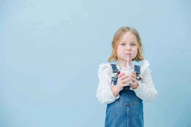 Photo of Cute little girl in overalls drinking milkshake over blue background