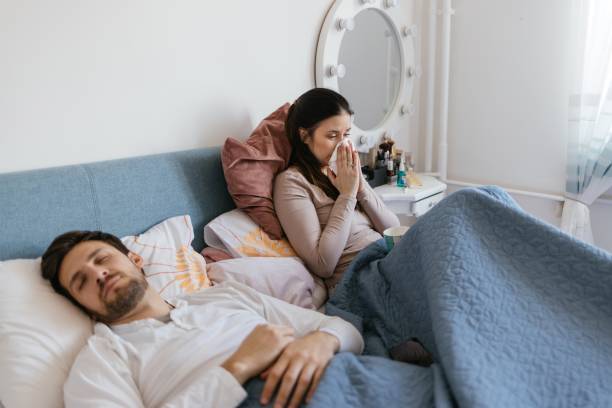 donna malata seduta sul letto e che soffia il naso mentre suo marito dorme - cold and flu flu virus bed couple foto e immagini stock