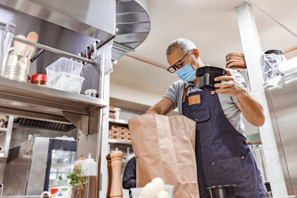 istock Mature professional chef in apron and protective mask preparing paper bag for takeaway delivery. 1305053549