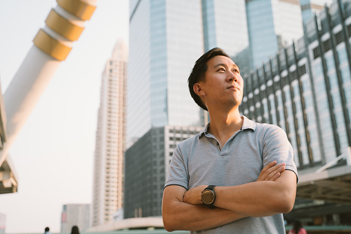 Young Asian man in polo shirt looking far forward to the future in high rise building exterior background