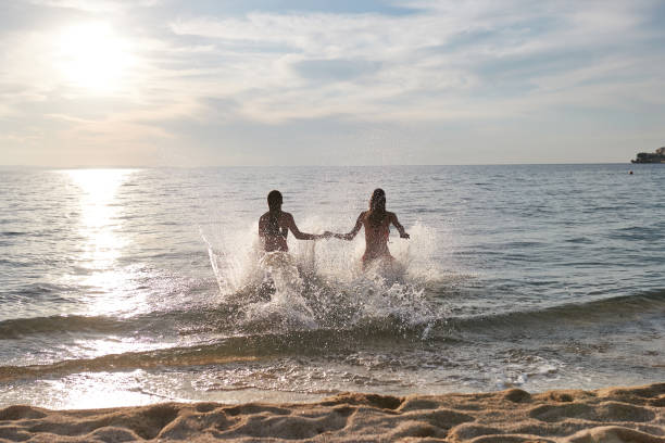 istock Two young women holding hands and splashing while running into the sea 1305056963