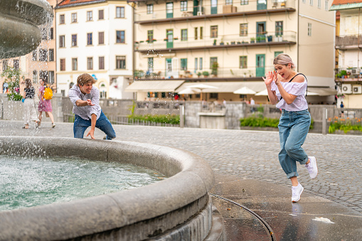 Young cherful couple splashing each other with fountain water in the city on a summer day.