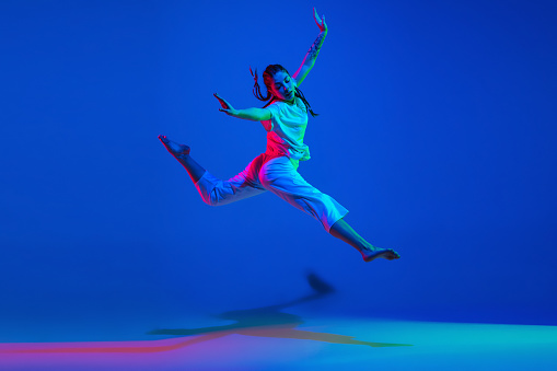 Flying. Sportive girl hip-hop dancer training in stylish white clothes on colorful background at dance hall in blue neon light. Youth culture, movement, fashion, action. Fashionable bright portrait.