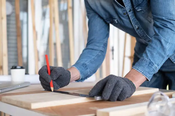 Photo of Professional carpenter man wearing glove using pencil and ruler to draw the line on the wood piece in the modern wood workplace. There are goggles. laptop and saw on the table. Furniture DIY concept