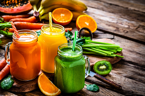 Healthy eating: fresh colored fruits and vegetables smoothies in Mason jars arranged on rustic wooden table. Fruits and vegetables like bananas, papaya, kiwi, spinach leaves, oranges and carrots are out of focus at background. High resolution 42Mp studio digital capture taken with Sony A7rII and Sony FE 90mm f2.8 macro G OSS lens