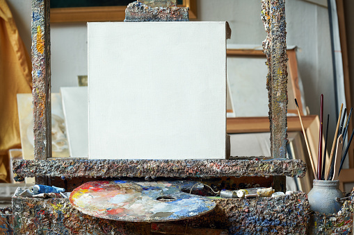 Artistic equipment in a artist studio: empty artist canvas on a wooden easel, paint tubes and paint brushes - used artistic paintbrushes for painting with oil or acrylic paints.