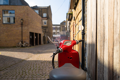 Color image depicting a retro-style red motorised scooter parked on a cobblestone city street in Notting Hill, London, UK.