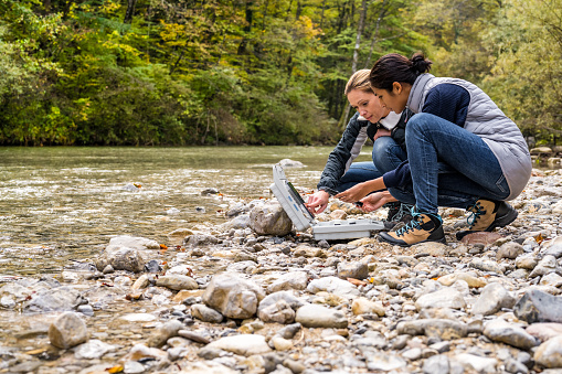 Vista de ángulo bajo de las investigadoras biológicas femeninas en cuclillas en la orilla del río y discutiendo datos medidos. photo