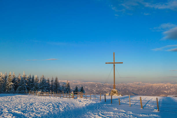 A huge wooden cross on top of Schoeckl, Austrian Alps during the morning golden hour. The peak is covered with powder snow. The mountain chains in the back are turned golden. Soft sunrise colors. una enorme cruz de madera en la parte superior de schoeckl, alpes austriacos durante la hora dorada de la mañana. el pico está cubierto de nieve en polvo. las cadenas de montaña en la parte posterior se vuelven doradas. colores suaves del amanecer.