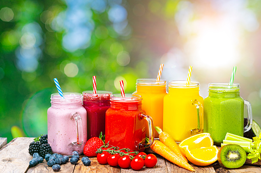 Healthy eating: fresh rainbow colored fruits and vegetables smoothies in Mason jars arranged side by side on rustic wooden table shot outdoors with defocused lush foliage at background. Fruits and vegetables like tomatoes, cucumber, oranges, strawberries, kiwi and carrots complete the composition. Copy space. High resolution 42Mp outdoors digital capture taken with Sony A7rII and Sony FE 90mm f2.8 macro G OSS lens