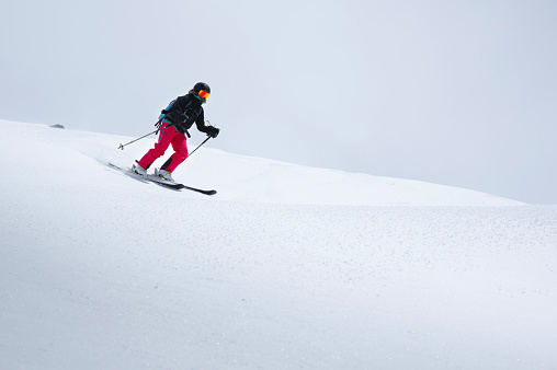 A freerider skier in red pants with a rockzack rides down a snowy slope in cloudy weather. Womens freeride sports background.