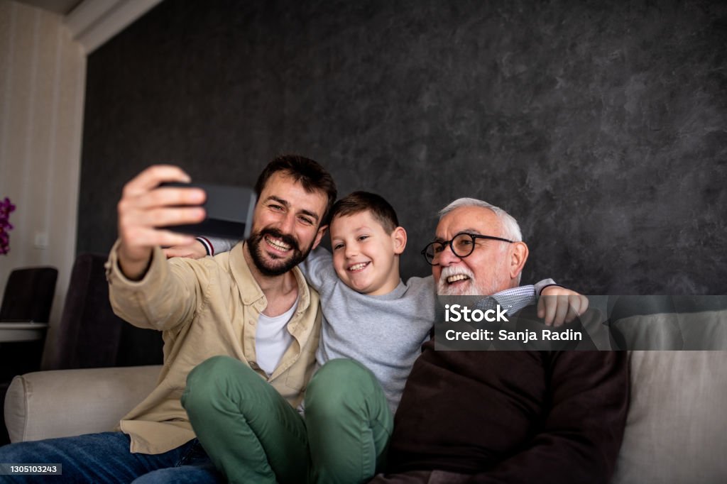 Smiling boys are sitting on sofa bed and taking selfie. A young adult man is holding his mobile phone and taking a selfie with his family. It is Father's day and grandson, father, and grandfather are enjoying their time together. Father's Day Stock Photo