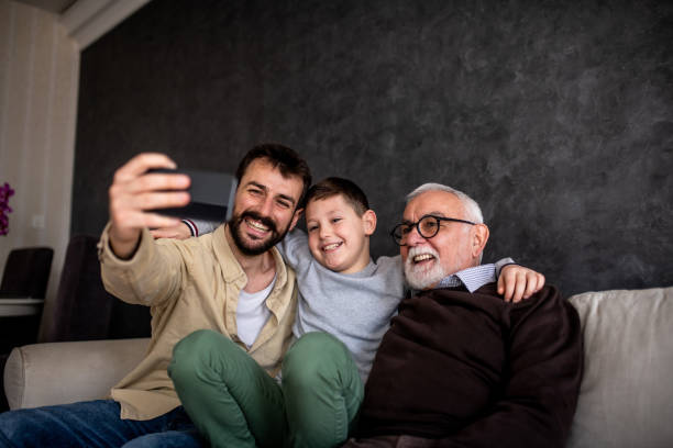 Smiling boys are sitting on sofa bed and taking selfie. A young adult man is holding his mobile phone and taking a selfie with his family. It is Father's day and grandson, father, and grandfather are enjoying their time together. father son grandfather fathers day stock pictures, royalty-free photos & images