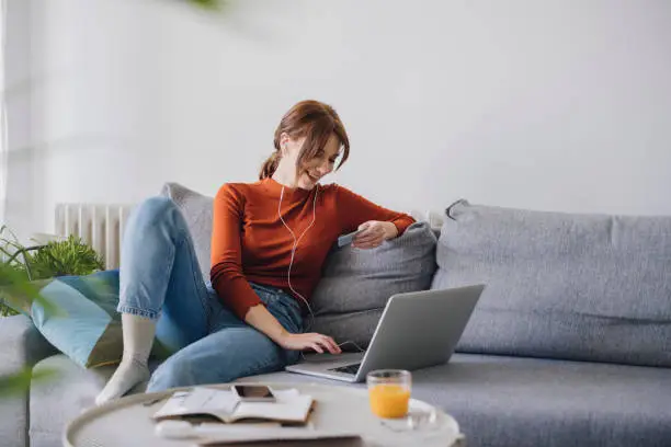 A beautiful young Caucasian businesswoman wearing a red long sleeve shirt and jeans holding a credit card and using a laptop for online shopping while sitting comfortably on a sofa at home.