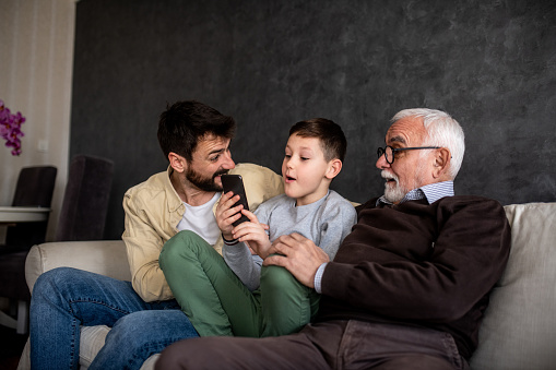 A young boy is holding a mobile phone while his father and grandfather are sitting by him, on the sofa bed. They are enjoying the weekend together.