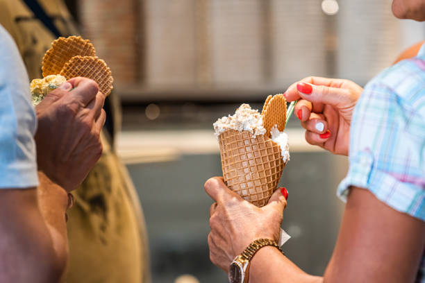 main de femme et d’homme retenant le cône blanc de gelato de crème glacée de vanille avec le fond et la cuillère flous de bokeh - ice cream parlor photos et images de collection