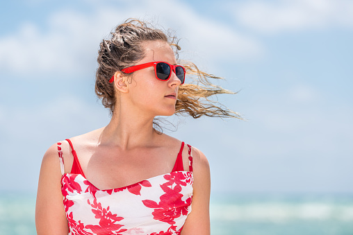 Portrait closeup of young woman hipster millennial in dress on beach during sunny day with red sunglasses in Miami, Florida with ocean in blurry blurred background