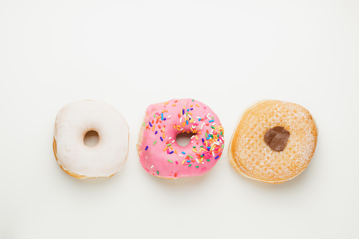 Overhead flat lay view of three fresh, colourful ring doughnuts in a row isolated on a plain white surface and background.
