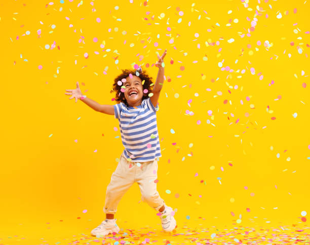 Excited African American kid jumping and catching confetti in studio Full body of joyful little black child with Afro hair in stylish clothes laughing and jumping while trying to catch colorful confetti against yellow background catching photos stock pictures, royalty-free photos & images