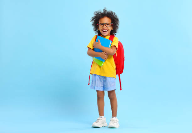 Cheerful little ethnic schoolchild with backpack and textbooks in studio Full body of positive little African American boy with curly hair in casual clothes and eyeglasses smiling while standing against blue background with school backpack and copybooks schoolboy stock pictures, royalty-free photos & images