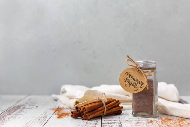 istock Jar of Cinnamon Sugar on a White Table with Gray Background; Bundle of Cinnamon Sticks and Ground Cinnamon on Table 1305130928