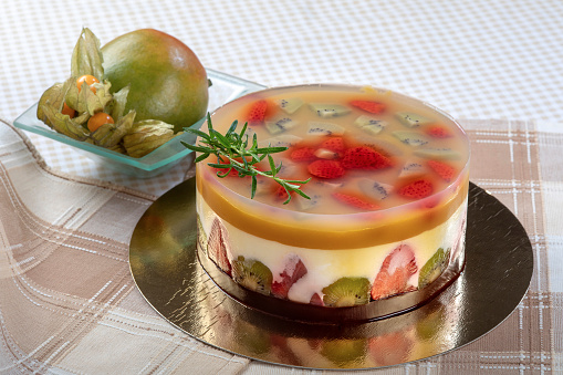 Refreshing cake with gelatin and fruit. The cake is filled with cream cheese and mango puree and decorated with rosemary. In the bowl next to the cake is mango and cape gooseberry.