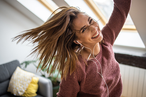 Cheerful young woman listening music on headphones and dancing in living room