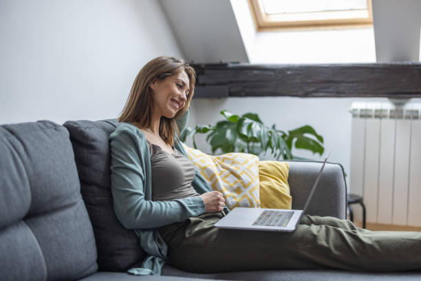istock Young smiling woman sitting on sofa and using laptop 1305135415