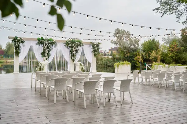 Place for wedding ceremony with wedding arch decorated with palm leaves, orchid flowers and floral peacocks, bulbs garland and white chairs outdoors, copy space