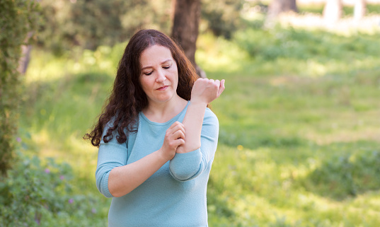 Woman scratching arm because it stings in a park with a green background