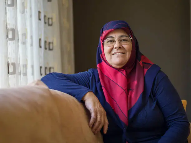 Muslim senior woman wearing a headscarf sitting on a sofa at home, daylight portrait