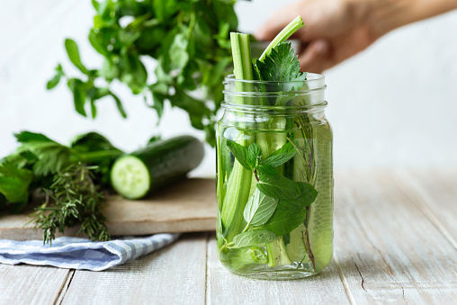Infused water bottle with an abundance cucumber slices, mint leafs, celery stalk and rosemary in on white wood. Hand of an unrecognizable person in the background is preparing the bottle with more green vegetables.